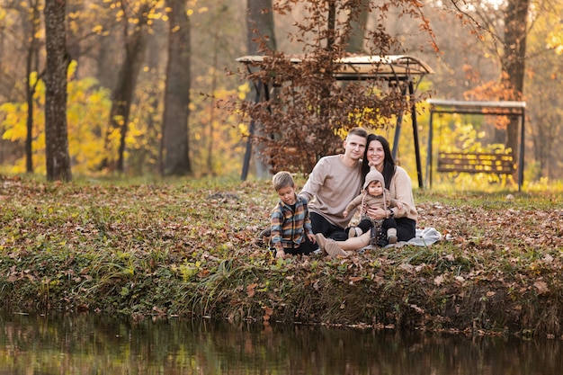 Jonge vader en moeder met babymeisje en zoontje plezier op deken in de buurt van vijver in herfst park