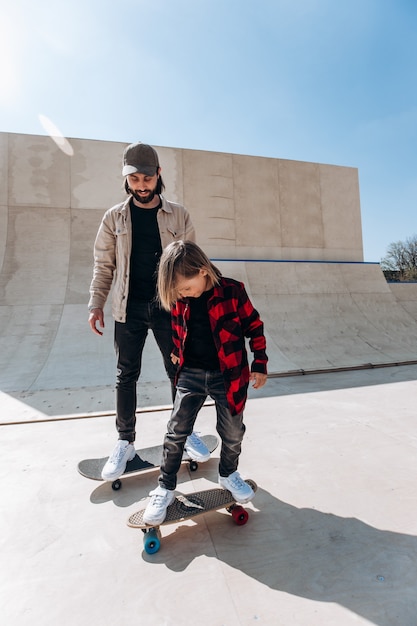 Jonge vader en zijn zoon rijden skateboards in een skatepark met glijbanen buiten op de zonnige dag.