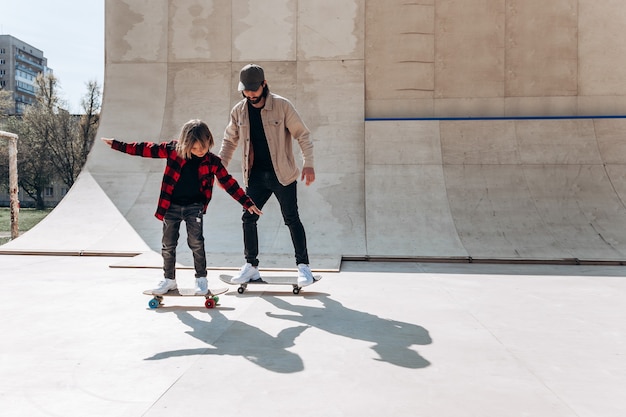 Jonge vader en zijn zoon rijden skateboards in een skatepark met glijbanen buiten op de zonnige dag.