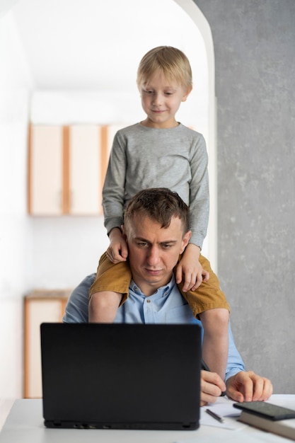 Jonge vader probeert thuis met de kinderen te werken. Zoon zit op zijn vaders schouders terwijl hij aan de laptop werkt.