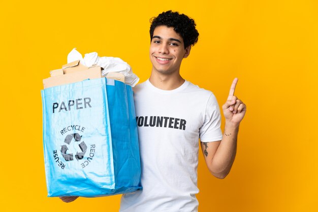 Jonge venezolaanse man die een recyclezak vol papier vasthoudt om te recyclen, toont en steekt een vinger op als teken van het beste