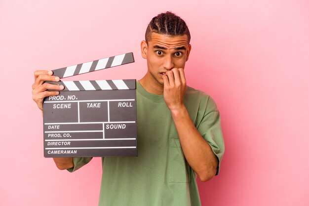 Jonge venezolaanse man met een filmklapper geïsoleerd op roze muur bijtende vingernagels, nerveus en erg angstig.