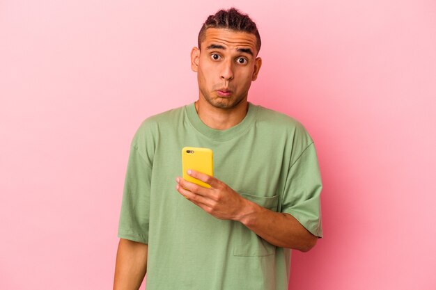Jonge venezolaanse man met een mobiele telefoon geïsoleerd op een roze muur haalt zijn schouders op en opent verwarde ogen.