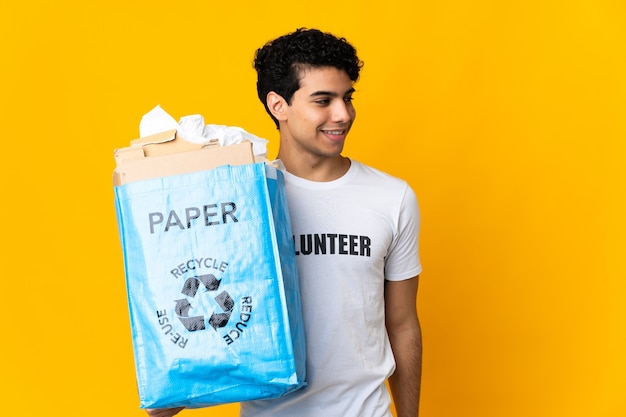 Jonge Venezolaanse man met een recyclezak vol papier om te recyclen, opzij kijkend en glimlachend