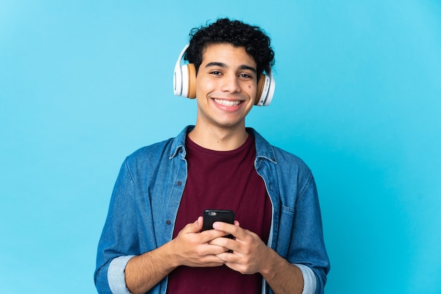 Jonge Venezolaanse man op blauwe muziek luisteren met een mobiel en naar voren kijken