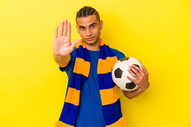 Jonge venezolaanse man voetbal kijken geïsoleerd op gele achtergrond permanent met uitgestrekte hand weergegeven: stopbord, waardoor u.