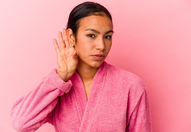 Jonge venezolaanse vrouw die een badjas draagt die op roze achtergrond wordt geïsoleerd en een roddel probeert te luisteren.