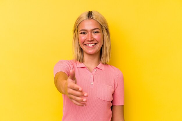 Jonge Venezolaanse vrouw die op gele muur wordt geïsoleerd die hand aan voorzijde in groetgebaar uitrekt