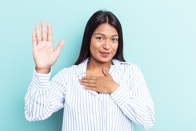 Jonge Venezolaanse vrouw geïsoleerd op een blauwe achtergrond die een eed aflegt, hand op de borst.