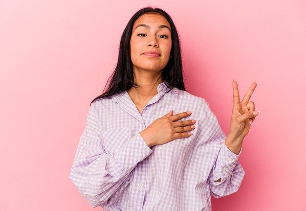 Jonge Venezolaanse vrouw geïsoleerd op roze achtergrond die een eed aflegt, hand op de borst.