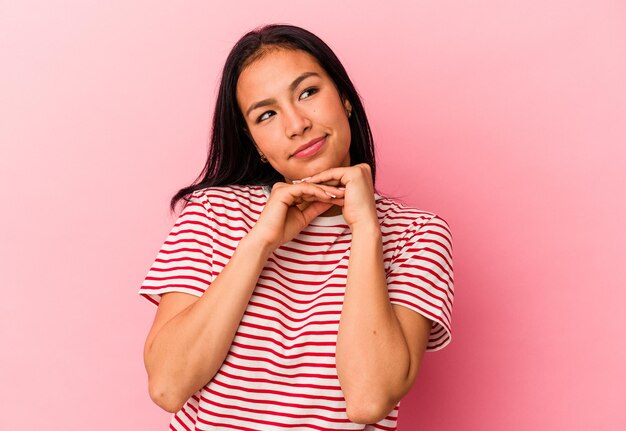 Jonge Venezolaanse vrouw geïsoleerd op roze achtergrond houdt handen onder de kin en kijkt vrolijk opzij