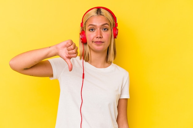 Jonge Venezolaanse vrouw luisteren muziek geïsoleerd op gele muur met een afkeer gebaar, duimen naar beneden. Meningsverschil concept.