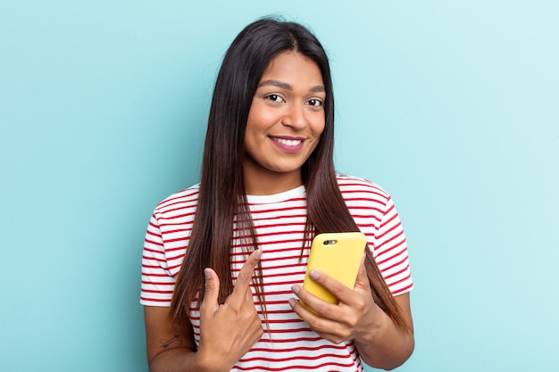 Jonge Venezolaanse vrouw met mobiele telefoon geïsoleerd op blauwe achtergrond wijzend met de vinger naar je alsof uitnodigend dichterbij komt.