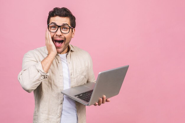 Jonge verbaasde geschokte bedrijfsmens die glazen dragen die computerlaptop met een glimlach werken die over roze muur wordt geïsoleerd.