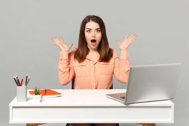 Jonge verbaasde vrouw in pastelkleren die mond open houden en handen spreiden, werken aan een bureau met pc-laptop geïsoleerd op een grijze achtergrond. Prestatie zakelijke carrière levensstijl concept. Bespotten kopie ruimte.