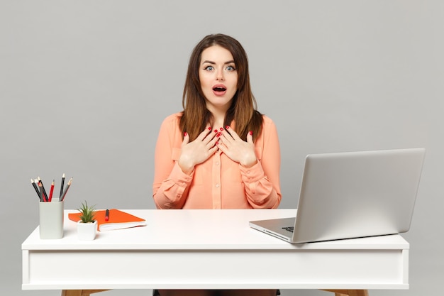 Jonge verbaasde vrouw in pastelkleren die mond open houden, handen op de borst houden, werken aan bureau met pc-laptop geïsoleerd op een grijze achtergrond. Prestatie zakelijke carrière concept. Bespotten kopie ruimte.