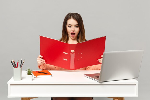 Jonge verbaasde vrouw in pastelkleren met rode map voor papieren document, werk aan bureau met pc-laptop geïsoleerd op een grijze achtergrond. Prestatie zakelijke carrière levensstijl concept. Bespotten kopie ruimte.