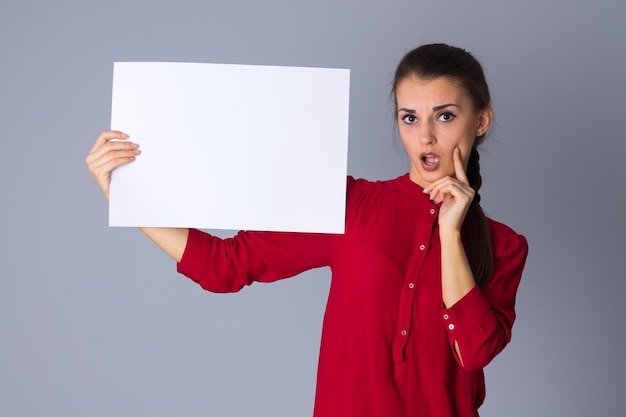 Jonge verbaasde vrouw in rode blouse met wit vel papier op grijze achtergrond in studio