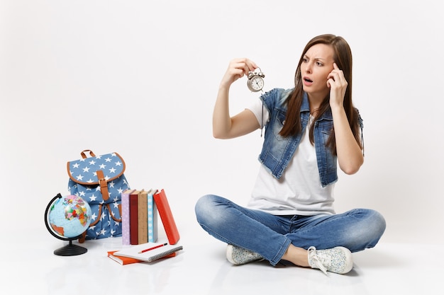 Jonge, verbijsterde, verbaasde vrouw die een wekker vasthoudt en de hand op de tempel houdt, zit in de buurt van de wereldbol, rugzak, schoolboeken geïsoleerd