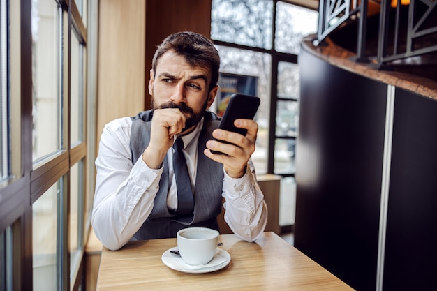 Jonge verdachte zakenman die in coffeeshop zit, slimme telefoon houdt en over e-mail denkt die hij heeft gelezen.