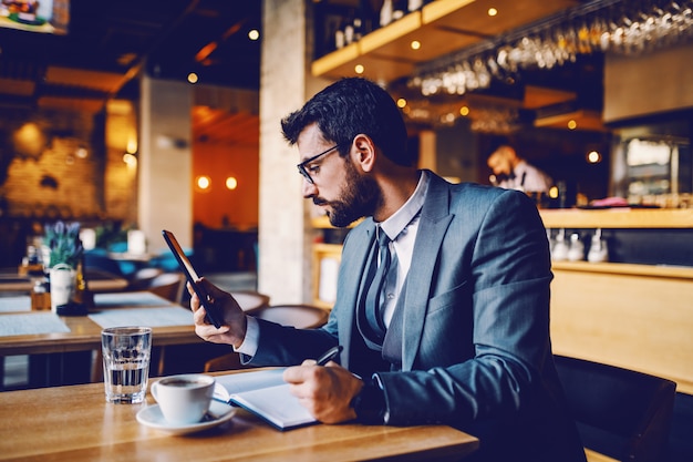 Jonge verfijnde kaukasische advocaat in pak met bril zitten in café, tablet kijken en belangrijke dingen schrijven over zaak in notitieblok.