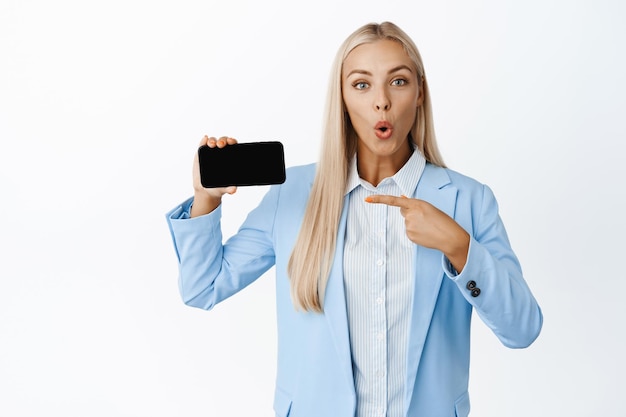 Jonge verkoopster die naar het horizontale smartphonescherm wijst en er verbaasd uitziet met een interessante app die op een witte achtergrond staat