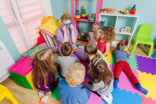 Jonge verloofde leraar zit met een groep kleuters op een vloer tijdens de les Leerproces kan leuk zijn Kamer met kleurrijke matten op een vloer en veelkleurige meubels