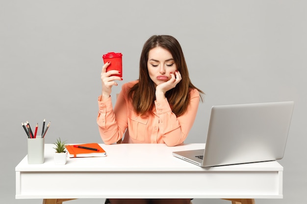 Jonge vermoeide vrouw die de ogen gesloten houdt en een papieren kopje koffie of thee vasthoudt, zit aan het bureau met een pc-laptop geïsoleerd op een grijze achtergrond. Prestatie zakelijke carrière levensstijl concept. Bespotten kopie ruimte.