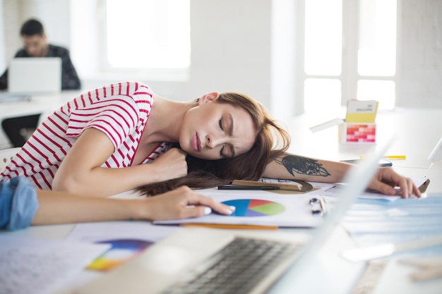 Jonge vermoeide vrouw in gestreept T-shirt die op het bureau slaapt terwijl ze tijd doorbrengt in een modern kantoor