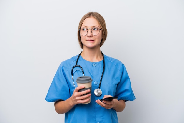 Jonge verpleegster arts vrouw geïsoleerd op een witte achtergrond met koffie om mee te nemen en een mobiel terwijl ze iets denkt