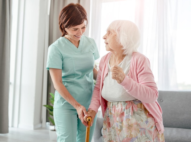 Jonge verpleegster helpt oudere vrouw thuis. Meisje in de gezondheidszorg geeft om senior vrouwelijke persoon binnenshuis