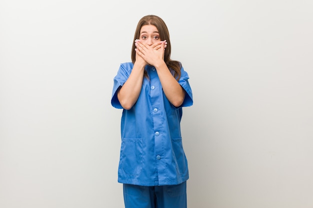 Jonge verpleegstersvrouw tegen een witte geschokte muur behandelend mond met handen.