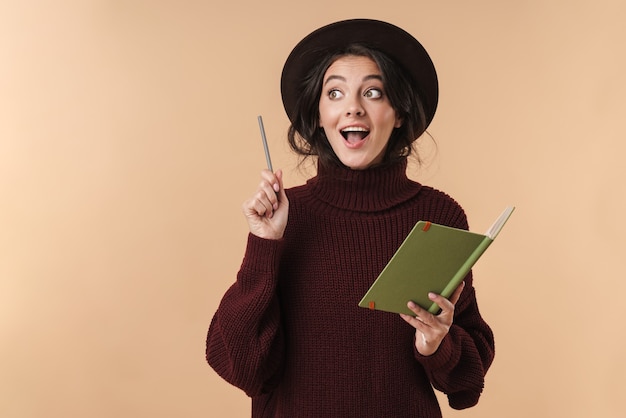 jonge verrast brunette vrouw geïsoleerd over beige muur muur schrijven van notities in notebook hebben een idee.