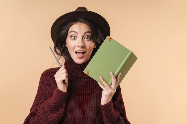 jonge verrast brunette vrouw geïsoleerd over beige muur muur schrijven van notities in notitieblok.