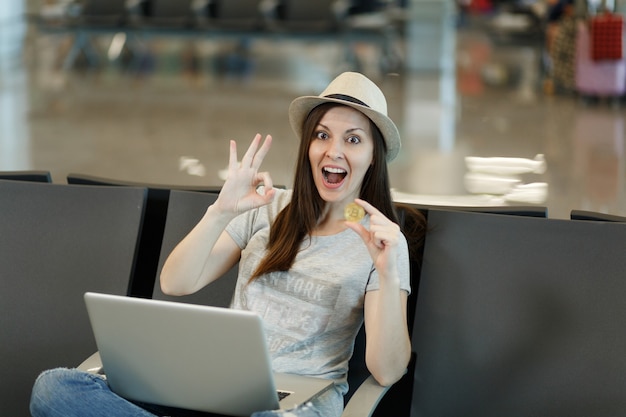 Jonge verraste reizigerstoerist met hoed zittend, werkend op laptop, bitcoin vasthoudend, OK-teken tonend, wachtend in lobbyhal op luchthaven