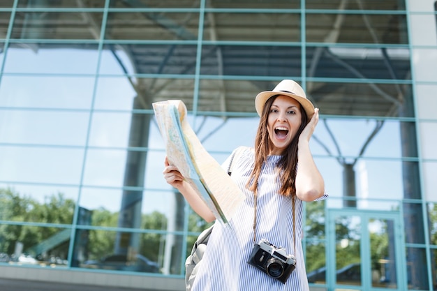 Jonge verraste reizigerstoeristenvrouw met retro vintage fotocamera, papieren kaart, zich vastklampend aan het hoofd op de internationale luchthaven