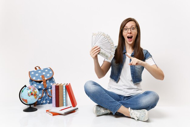 Jonge verraste vrouw student wijzende wijsvinger op bundel veel dollars, contant geld zittend in de buurt van globe, rugzak, boeken geïsoleerd