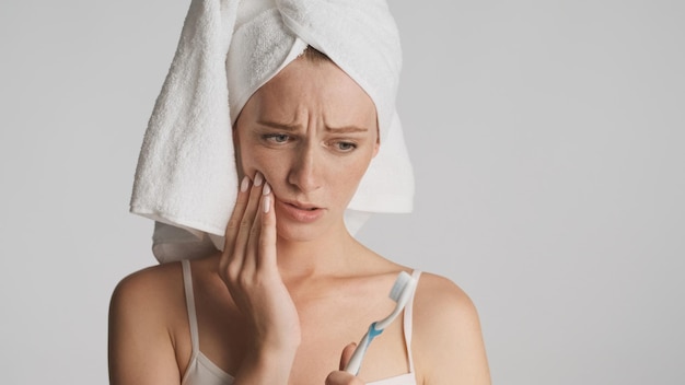 Jonge verstoorde vrouw met handdoek op hoofd die droevig op tandenborstel kijkt die op witte achtergrond wordt geïsoleerd