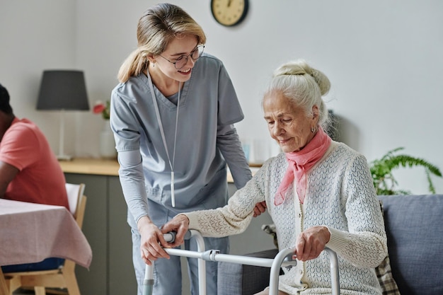 Foto jonge verzorger die om senior vrouw in verpleeghuis geeft, helpt haar om te staan met behulp van een rollator