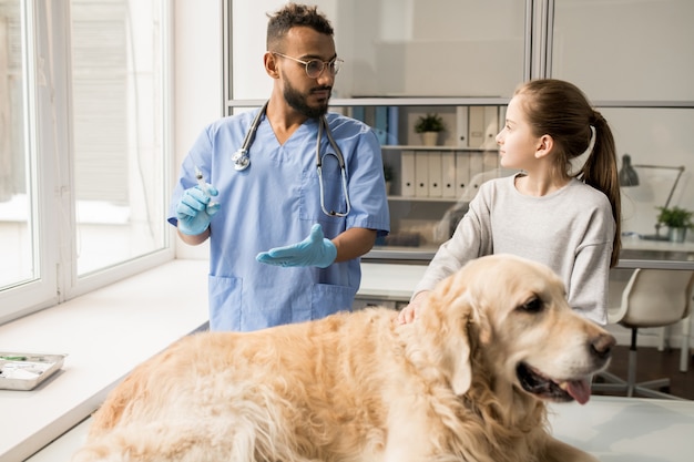 Jonge veterinaire professional in handschoenen en uniform spuit met geneeskunde voorbereiden en weinig eigenaar van labrador hond raadplegen