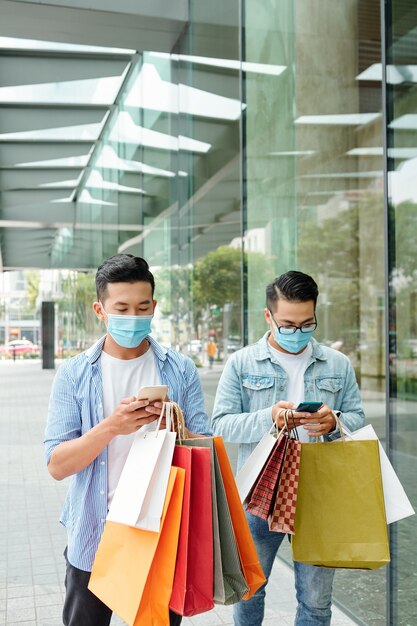 Jonge Vietnamese mannen in medische maskers die met boodschappentassen op straat lopen en vrienden sms'en of meldingen controleren