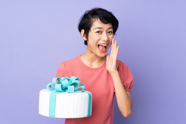 Jonge Vietnamese vrouw die met kort haar een grote cake over purpere muur houdt die met wijd open mond schreeuwt