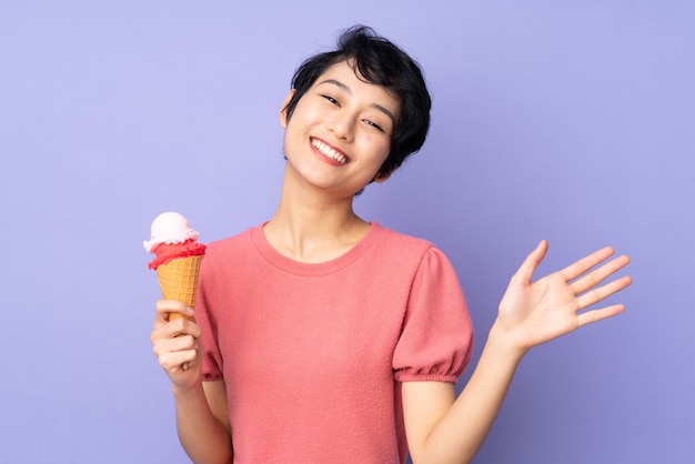 Jonge Vietnamese vrouw die met kort haar een kornetroomijs over purpere muur houden die met hand met gelukkige uitdrukking groeten