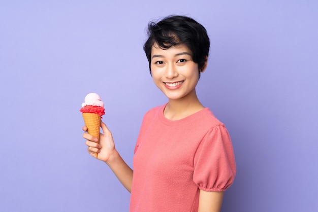 Jonge vietnamese vrouw die met kort haar een kornetroomijs over purpere muur houdt die veel glimlacht