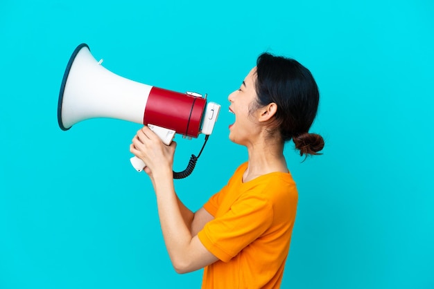 Jonge Vietnamese vrouw geïsoleerd op blauwe achtergrond die door een megafoon schreeuwt om iets in zijpositie aan te kondigen