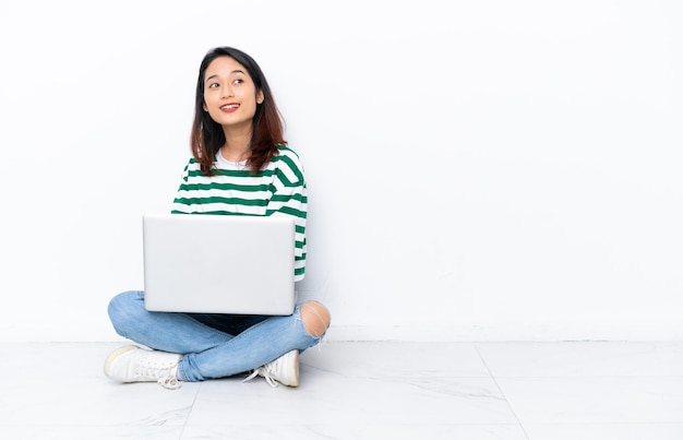 Jonge Vietnamese vrouw met een laptop zittend op de vloer geïsoleerd op een witte muur met gekruiste armen en happy