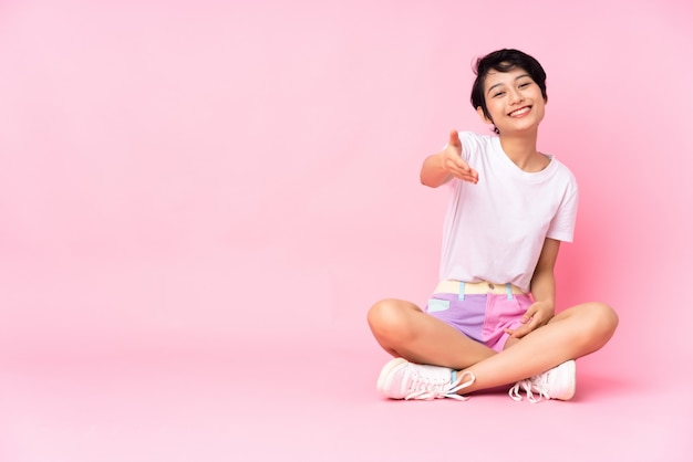 Jonge Vietnamese vrouw met kort haar zittend op de vloer over geïsoleerde roze muur handen schudden voor het sluiten van een goede deal