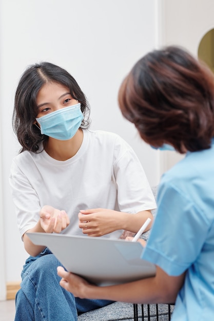 Jonge Vietnamese vrouw met medisch masker in gesprek met verpleegkundige van de tandheelkundige kliniek die het document vult