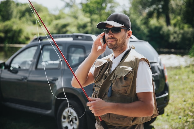 Jonge visser die op meer of rivier vist. Drukke man hengel vasthouden en praten over smartphone. Sta alleen bij de auto tijdens het sporten.