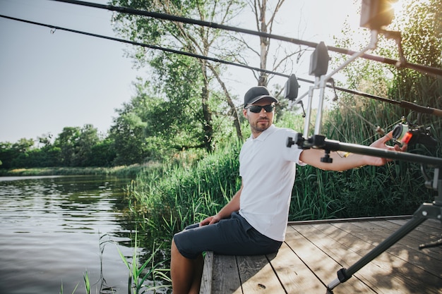 Jonge visser die op meer of rivier vist. kerel zit alleen bij de waterpier en kijkt terug op vishengels. pas ze aan voor waterjacht. visser benen in water houden.